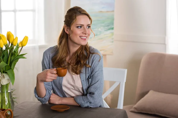 Sonriente joven mujer sosteniendo una taza de café y mirando a casa - foto de stock