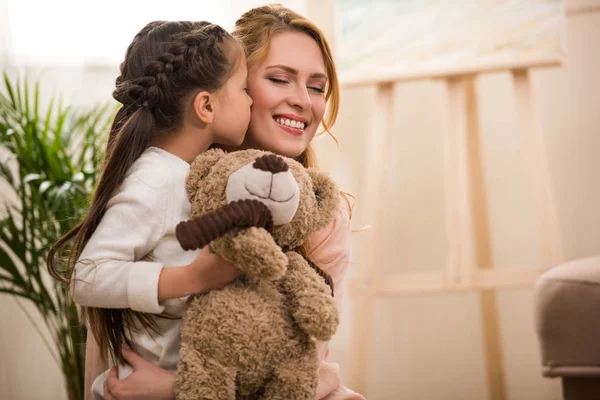 Cute little child holding teddy bear and kissing happy mother at home — Stock Photo