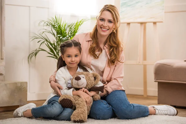 Glückliche Mutter und kleine Tochter mit Teddybär sitzen zusammen und lächeln zu Hause in die Kamera — Stockfoto