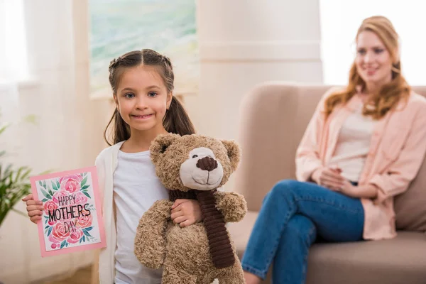 Adorable enfant tenant ours en peluche et joyeux jour des mères carte de vœux tandis que la mère assis derrière — Photo de stock