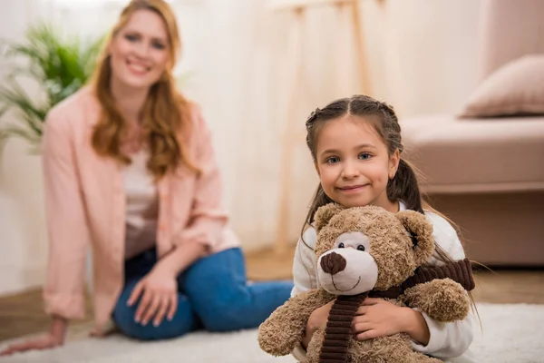 Adorable little child holding teddy bear and smiling at camera while mother sitting behind at home — Stock Photo