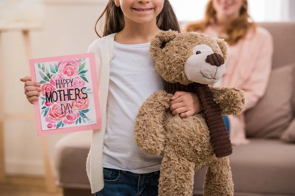 Recortado disparo de niño feliz sosteniendo osito de peluche y feliz madre día tarjeta de felicitación - foto de stock