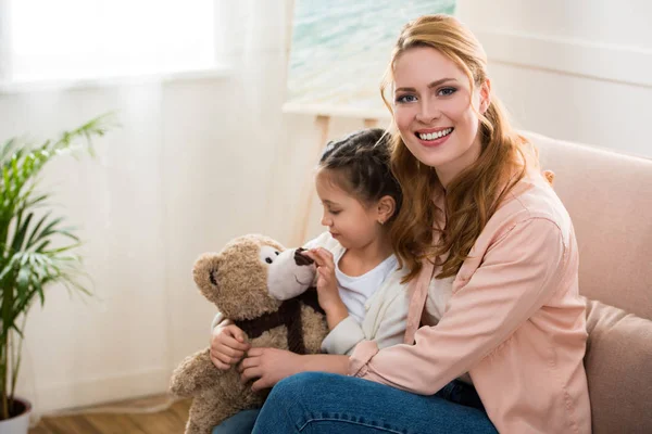 Happy young woman smiling at camera while daughter playing with teddy bear — Stock Photo