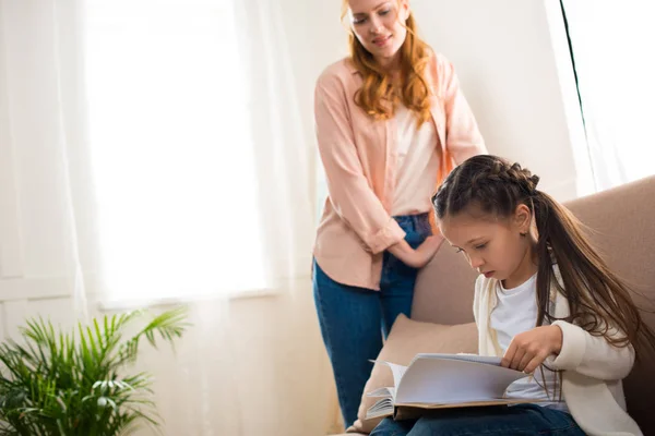 Mère heureuse regardant petite fille lecture livre à la maison — Photo de stock