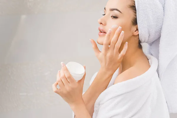 Belle femme en peignoir avec serviette sur la tête appliquant crème visage — Photo de stock