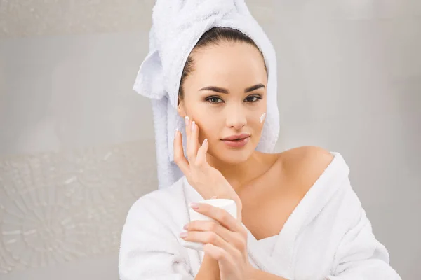 Portrait de belle femme avec serviette sur la tête tenant crème visage — Photo de stock