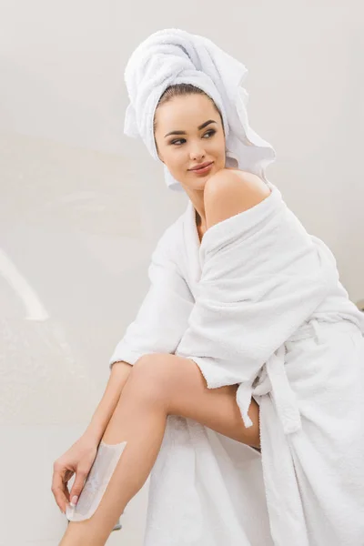 Belle femme en peignoir avec serviette sur la tête faisant épilation à la cire à la maison — Photo de stock