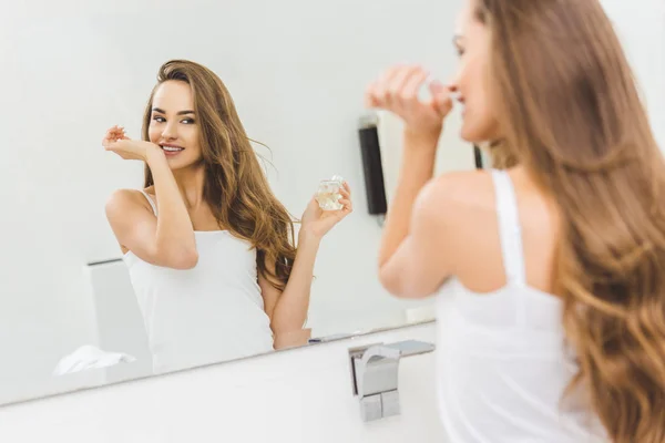 Mirror reflection of smiling woman smelling perfume on hand in bathroom — Stock Photo