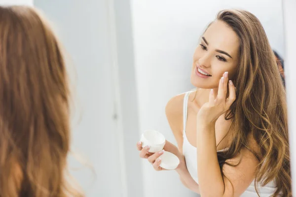 Mirror reflection of beautiful smiling woman applying face cream — Stock Photo