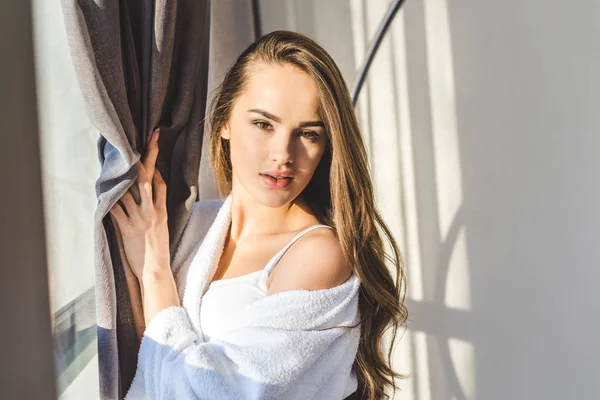 Portrait of pretty young woman in bathrobe standing at window at home — Stock Photo