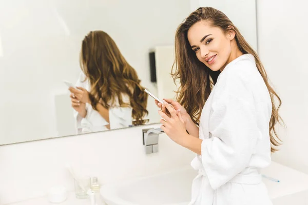 Portrait of smiling woman in bathrobe with smartphone in hands in bathroom — Stock Photo