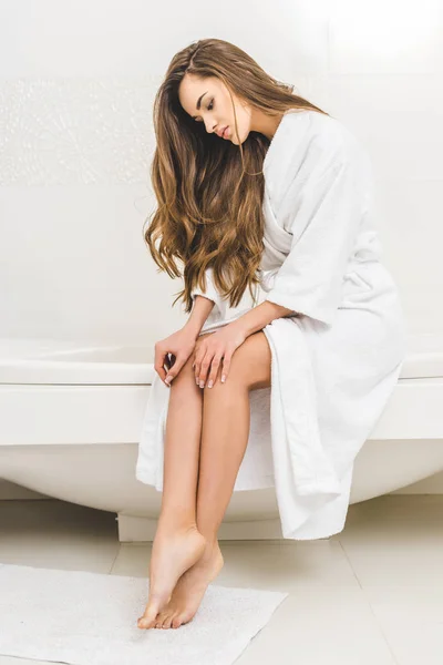 Mujer joven en albornoz sentado en el tubo de baño en casa - foto de stock
