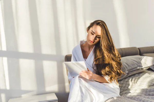 Ritratto di giovane donna in accappatoio con tablet seduta sul letto di casa — Foto stock