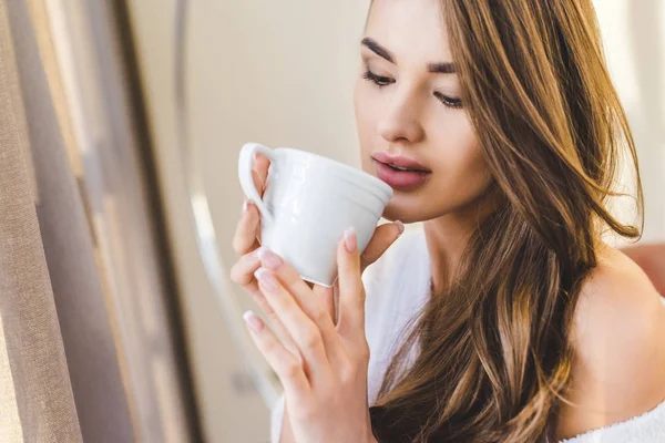 Portrait of beautiful young woman in bathrobe with cup of coffee at home — Stock Photo