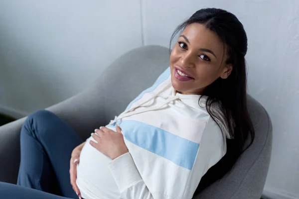 Feliz mulher grávida afro-americana sentada em poltrona e olhando para a câmera — Fotografia de Stock