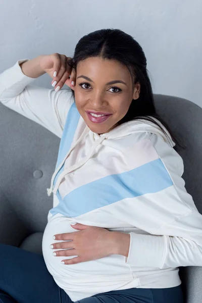 Africano americano embarazada mujer sentado en sillón y mirando a cámara - foto de stock