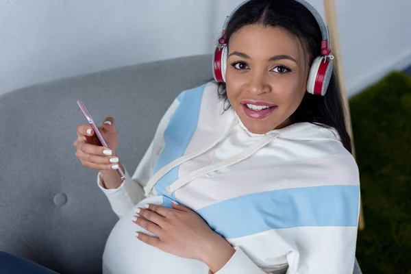 Mujer embarazada afroamericana sonriente escuchando música con auriculares y teléfono inteligente - foto de stock