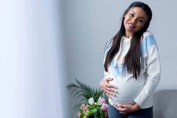 Happy african american pregnant woman looking at her belly while standing at window — Stock Photo