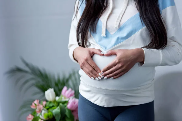 Recortado vista de embarazada chica haciendo símbolo del corazón con las manos en su vientre - foto de stock