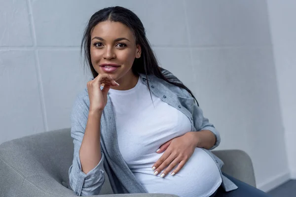 Lächelnde afrikanisch-amerikanische schwangere Frau blickt in die Kamera, während sie auf einem Sessel sitzt — Stockfoto