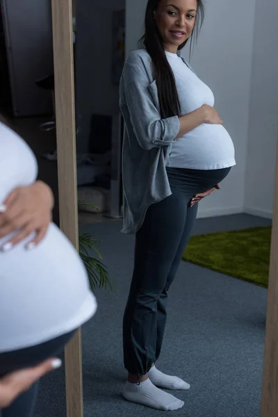 Mulher grávida afro-americana olhando para a barriga no espelho — Fotografia de Stock