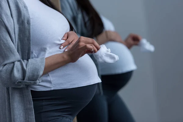 Vista recortada de la mujer embarazada sosteniendo pequeños calcetines en el espejo - foto de stock