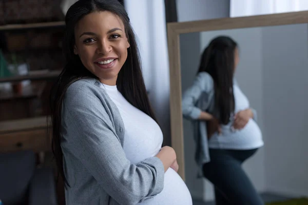 Cheerful african american pregnant woman standing at mirror — Stock Photo