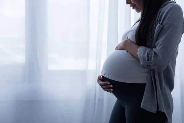Обрезанный вид беременной женщины, прикасающейся к животу, стоя у окна — стоковое фото