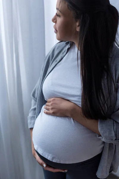 Mujer embarazada afroamericana tocando su vientre mientras mira a la ventana - foto de stock