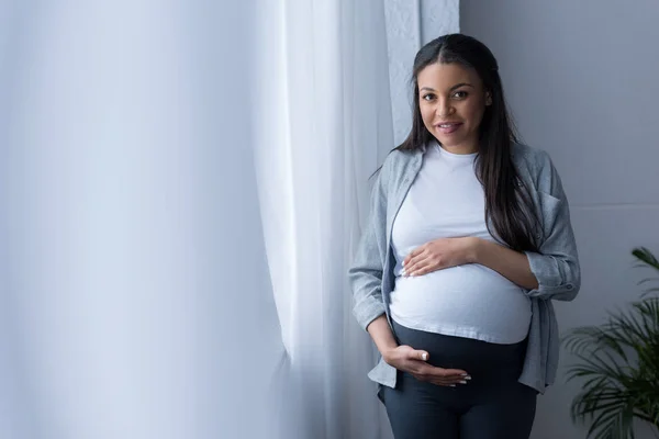 Smiling african american pregnant woman touching her belly while standing at window — Stock Photo