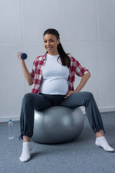 Mulher grávida afro-americana sentada em forma de bola e treinamento com halteres — Fotografia de Stock