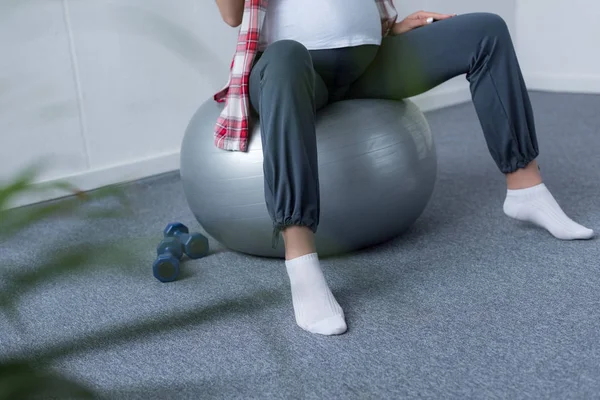 Обрезанный вид беременной женщины, сидящей на фитнес-мяч с гантелями рядом — стоковое фото