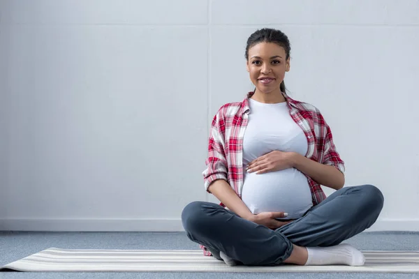 Mujer embarazada afroamericana sentada en esterilla de yoga - foto de stock
