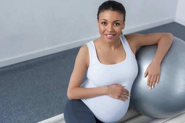 Fröhliche afrikanisch-amerikanische schwangere Frau sitzt mit fittem Ball und blickt in die Kamera — Stockfoto