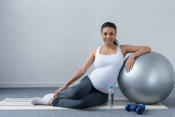Femme enceinte afro-américaine souriante assise avec balle en forme avec de l'eau et des haltères — Photo de stock