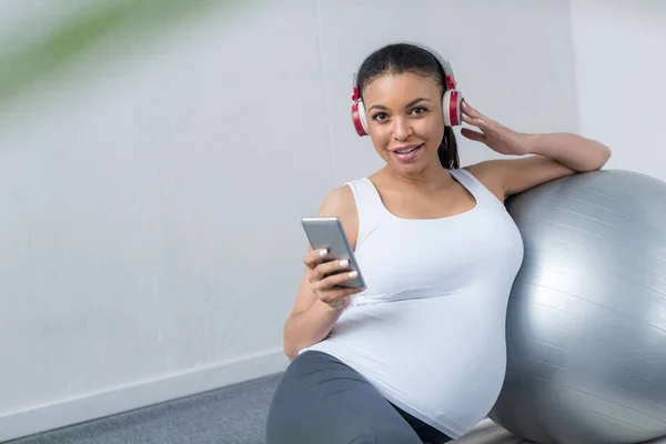 African american pregnant woman listening music with headphones and smartphone while sitting with fit ball — Stock Photo