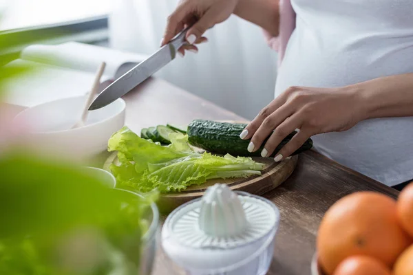 Cropped view of pregnant woman cutting cucumber and salad at kitchen — Stock Photo