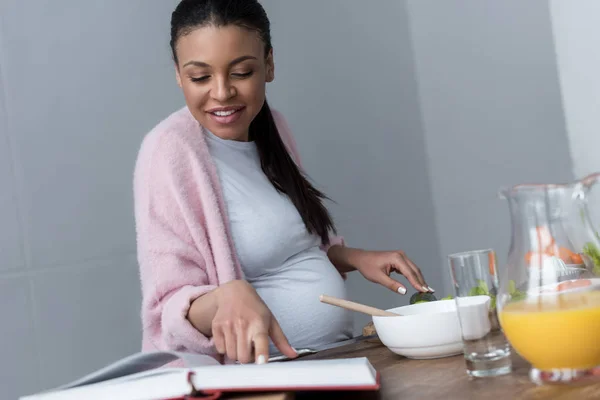 Smiling african american pregnant woman cooking and reading cookbook — Stock Photo