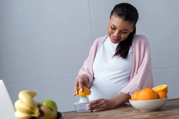 Happy african american pregnant woman making orange juice with squeezer — Stock Photo
