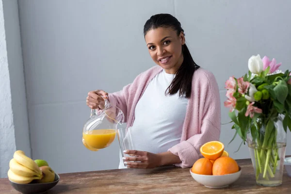 Afro-américaine enceinte verser du jus à la cuisine avec des fruits frais — Photo de stock