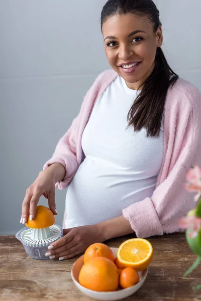 Hermosa sonriente africana americana embarazada mujer haciendo jugo de naranja con exprimidor - foto de stock