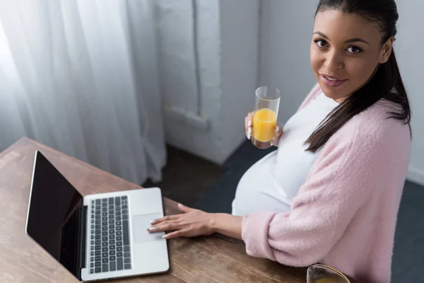 Mujer embarazada afroamericana con jugo de naranja utilizando el ordenador portátil - foto de stock