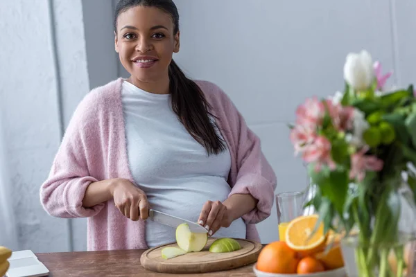 Afro-américaine enceinte coupe pomme à la cuisine — Photo de stock