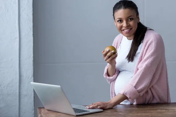 Mulher grávida afro-americana com maçã usando laptop — Fotografia de Stock