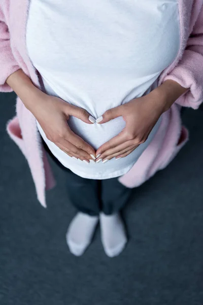 Vista recortada de la mujer embarazada haciendo símbolo del corazón con las manos en su vientre - foto de stock
