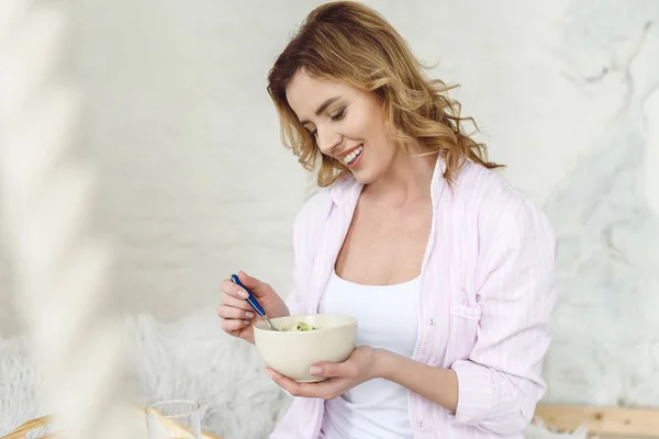 Happy blonde woman in pajamas holding bowl with breakfast — Stock Photo