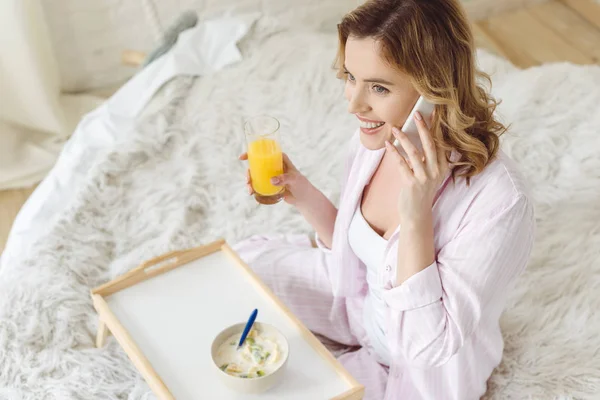 Веселая женщина в пижаме разговаривает по смартфону, сидя на кровати с завтраком на подносе — стоковое фото