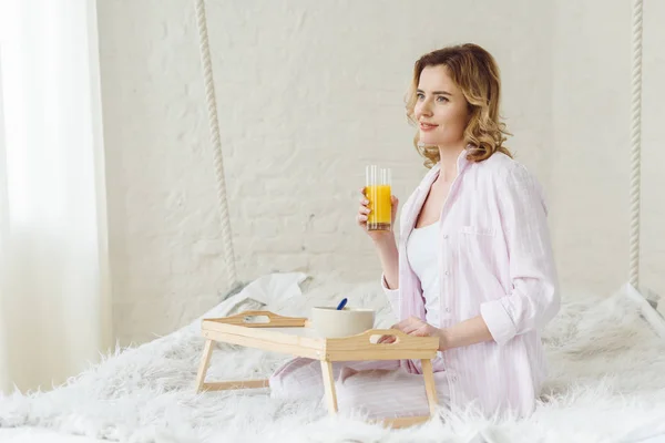 Femme blonde en pyjama assis sur le lit avec du jus d'orange et petit déjeuner sur le plateau — Photo de stock