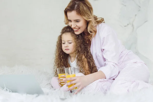 Mother and curly daughter with glasses of orange juice using laptop in bed — Stock Photo