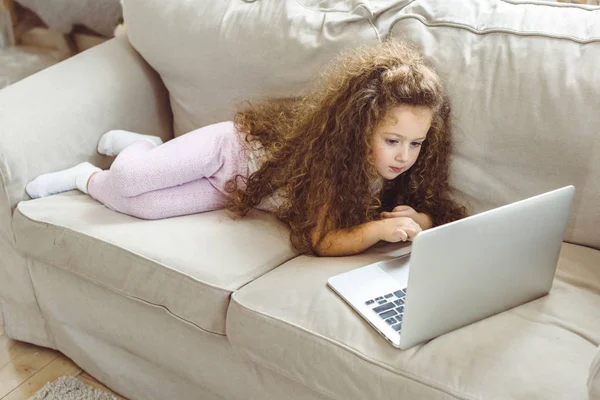 Adorabile bambino riccio utilizzando il computer portatile e sdraiato sul divano — Foto stock